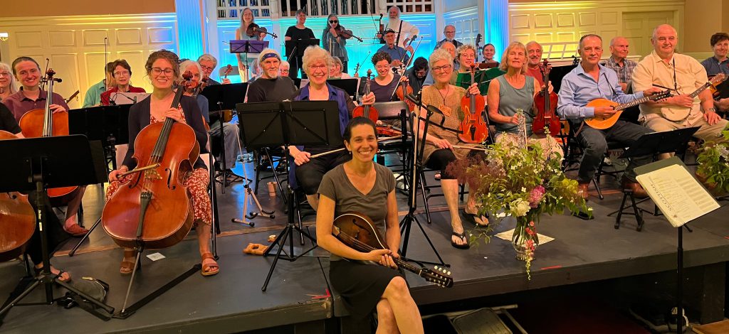 Annika sitting on the front of the stage with a smiling orchestra behind her